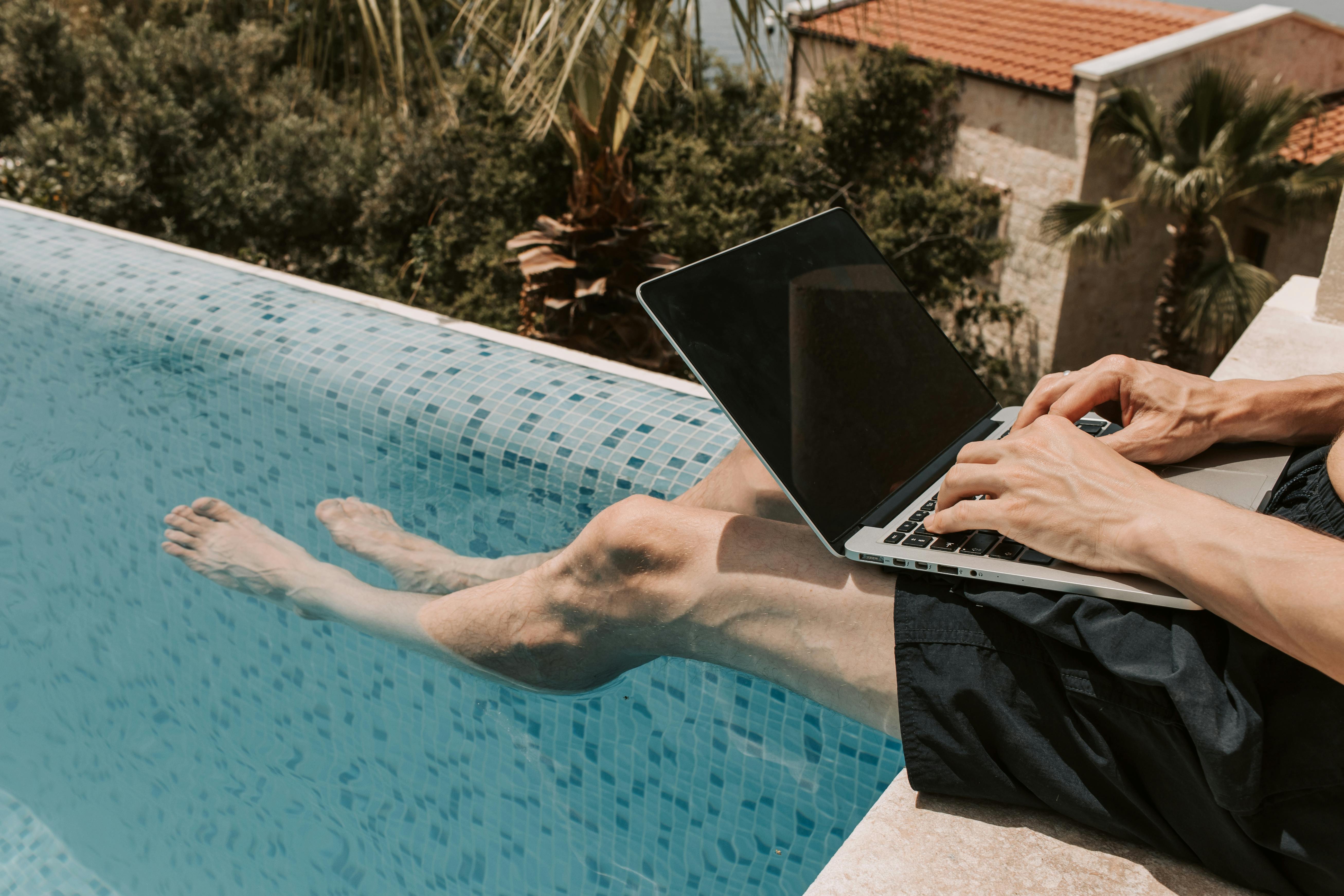 Man with computer and his legs are dangling in a swimming pool